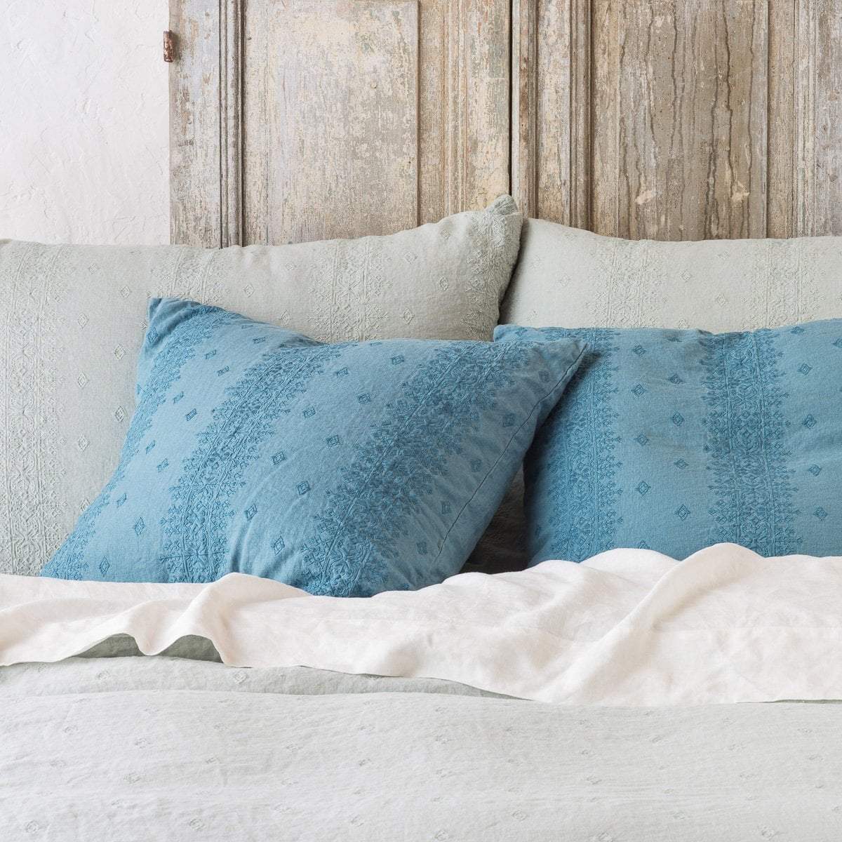 : Eucalyptus and cenote Ines shams leaning upright against a distressed wood headboard on light toned bedding - cropped head-on view.