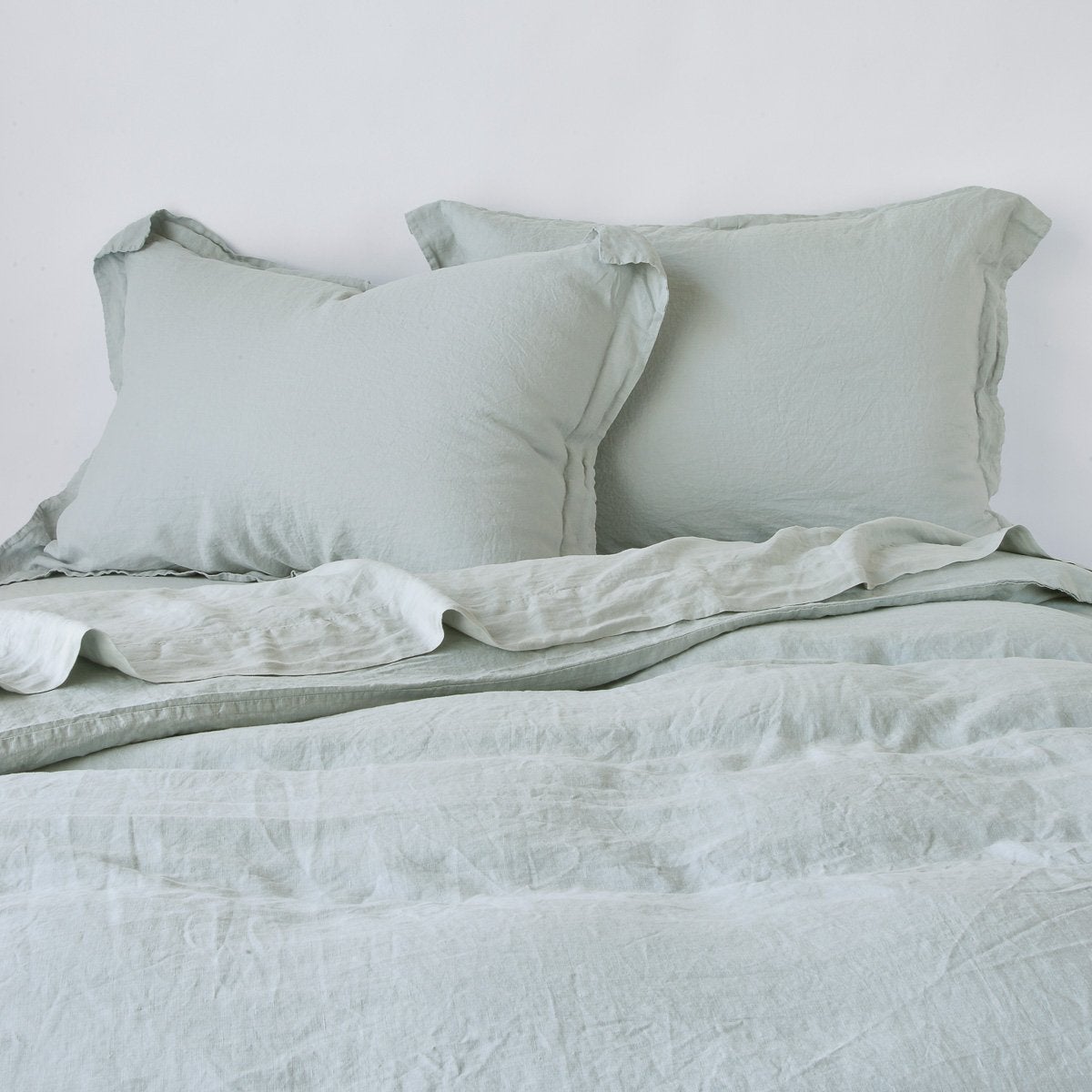 : Linen duvet cover in eucalyptus, partially folded back on a bed with matching sheets and shams on a plain background - cropped end of bed view.