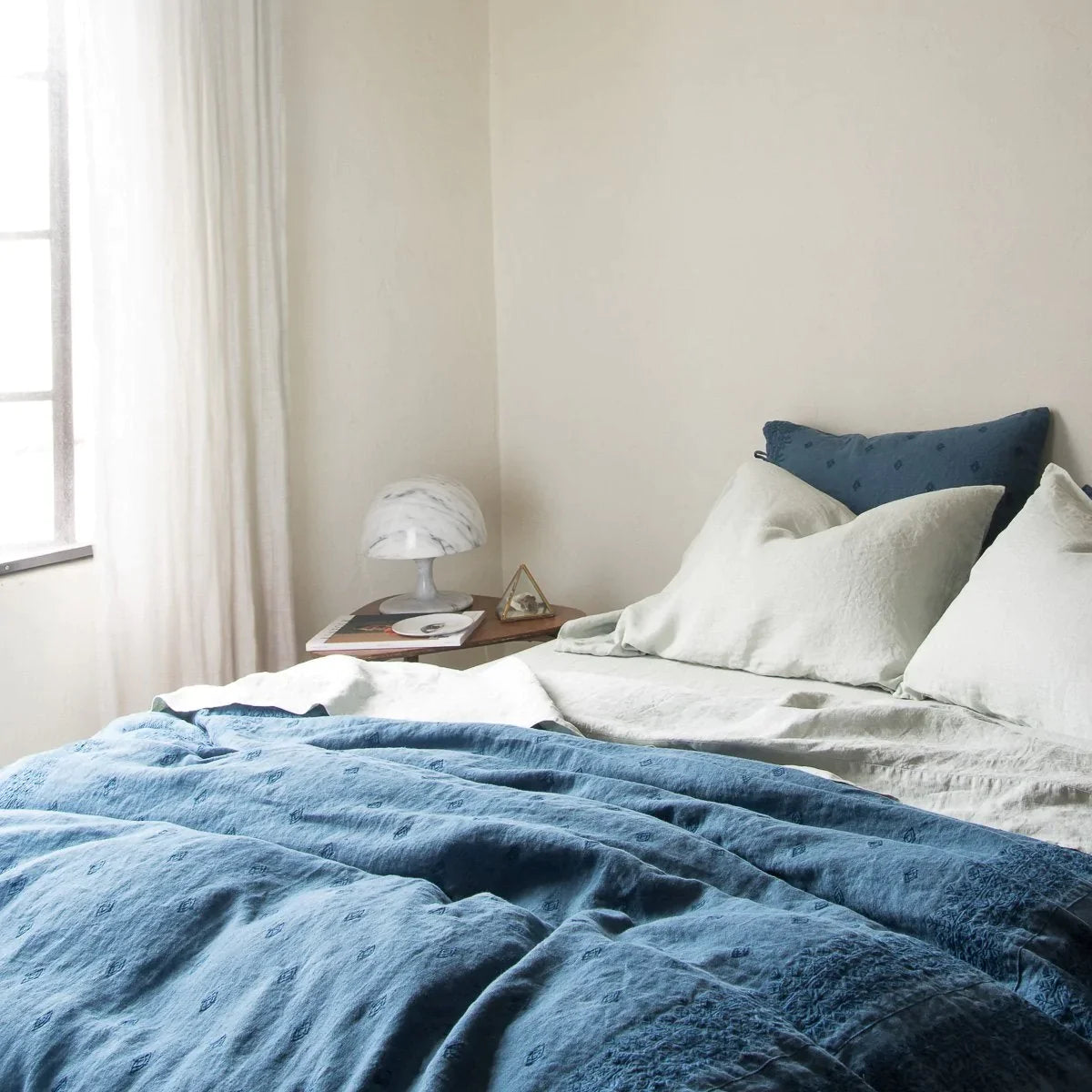 : Linen sheeting in eucalyptus, rumpled with duvet and shams in midnight embroidered linen - cropped three-quarter angle.