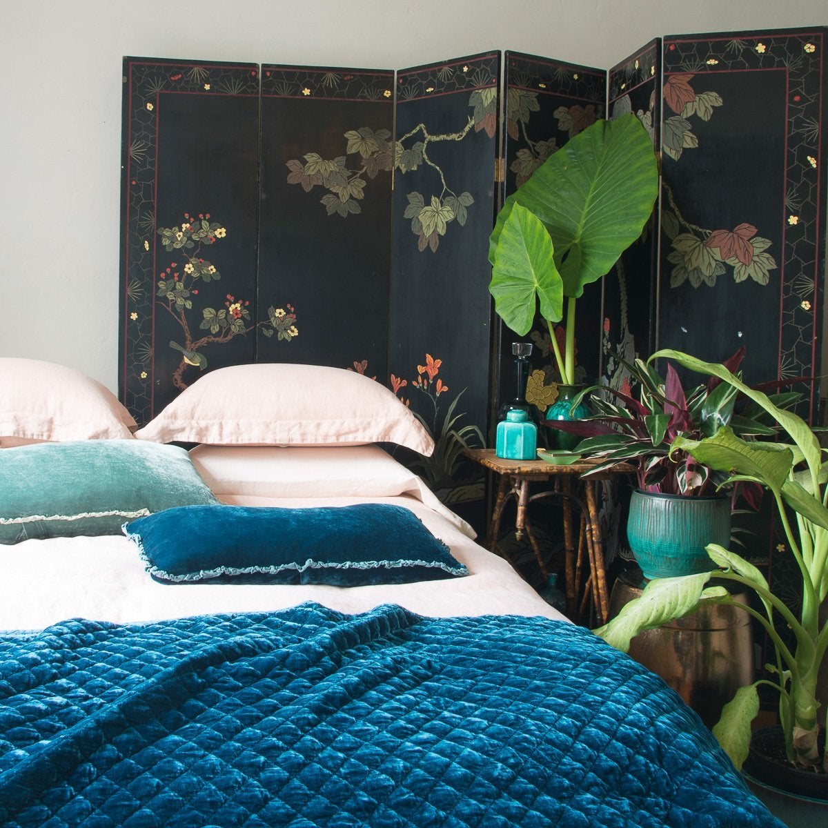 : Silk velvet quilted coverlet in cenote draped over the end of a pearl bed. Dark floral wall panel, blue-green ceramic vases, and big green plants complement the rich silk velvet - cropped three-quarter angle.